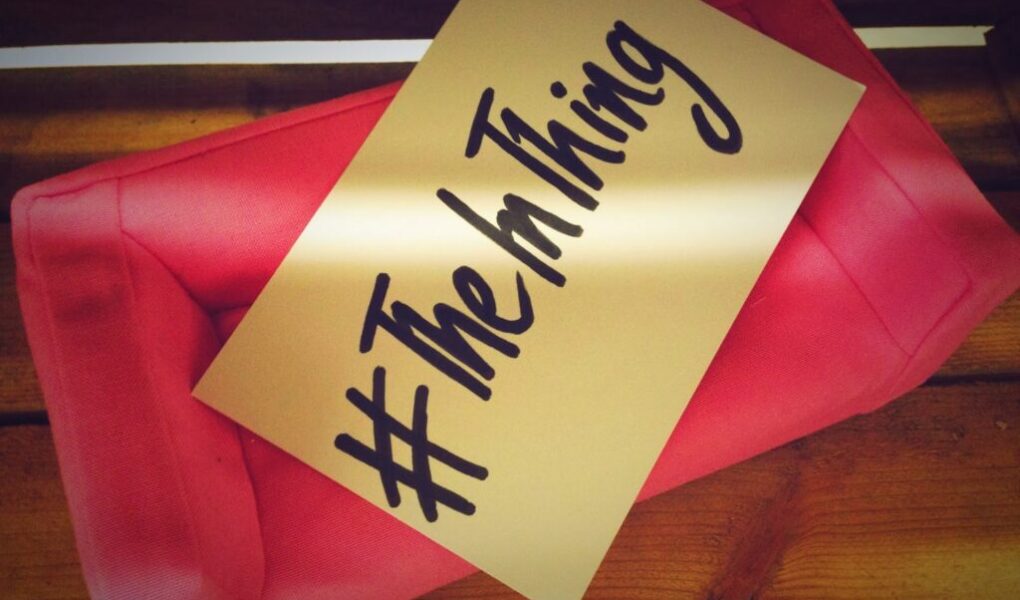 A pink fabric clutch bag sits on a wooden surface. Resting on the bag is a beige card with bold, black handwritten text that reads “#TheInThing.” The image is warm-toned, with soft lighting creating a cosy atmosphere.