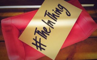 A pink fabric clutch bag sits on a wooden surface. Resting on the bag is a beige card with bold, black handwritten text that reads “#TheInThing.” The image is warm-toned, with soft lighting creating a cosy atmosphere.