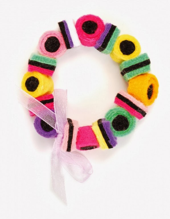 A circular bracelet made from colourful felt pieces resembling liquorice allsorts sweets, arranged in alternating colours. The bracelet is fastened with a light pink sheer ribbon tied in a bow, all set against a white background.