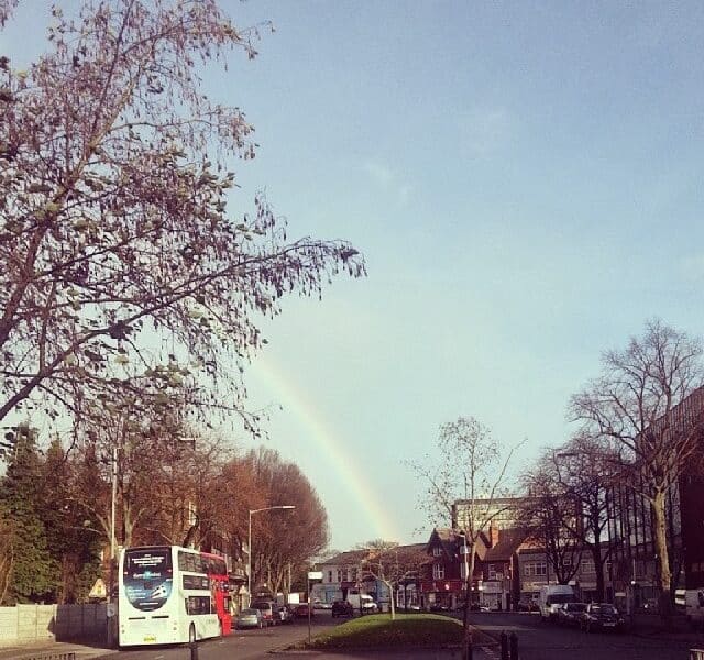 A street scene shows a double-decker bus driving away, trees lining both sides of the road, and buildings in the background. A faint rainbow arches over the houses under a mostly clear sky, with some bare branches visible in the foreground.