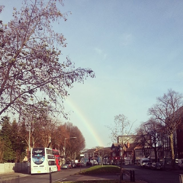 A street scene shows a double-decker bus driving away, trees lining both sides of the road, and buildings in the background. A faint rainbow arches over the houses under a mostly clear sky, with some bare branches visible in the foreground.