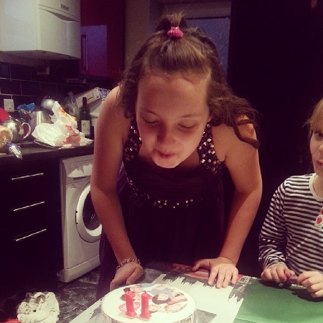 A young girl in a sleeveless dress leans over a small round birthday cake, blowing out two pink candles. Another child in a striped shirt sits beside her. They are in a kitchen, with various items and appliances visible in the background.