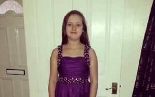 A young girl stands indoors in front of a white door, wearing a sleeveless purple dress with decorative embellishments on the straps and waist. She has straight brown hair and smiles slightly. A closed door with a letterbox is visible in the background.
