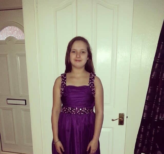 A young girl stands indoors in front of a white door, wearing a sleeveless purple dress with decorative embellishments on the straps and waist. She has straight brown hair and smiles slightly. A closed door with a letterbox is visible in the background.
