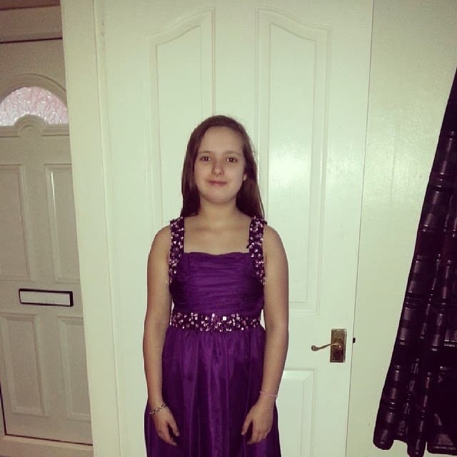A young girl stands indoors in front of a white door, wearing a sleeveless purple dress with decorative embellishments on the straps and waist. She has straight brown hair and smiles slightly. A closed door with a letterbox is visible in the background.