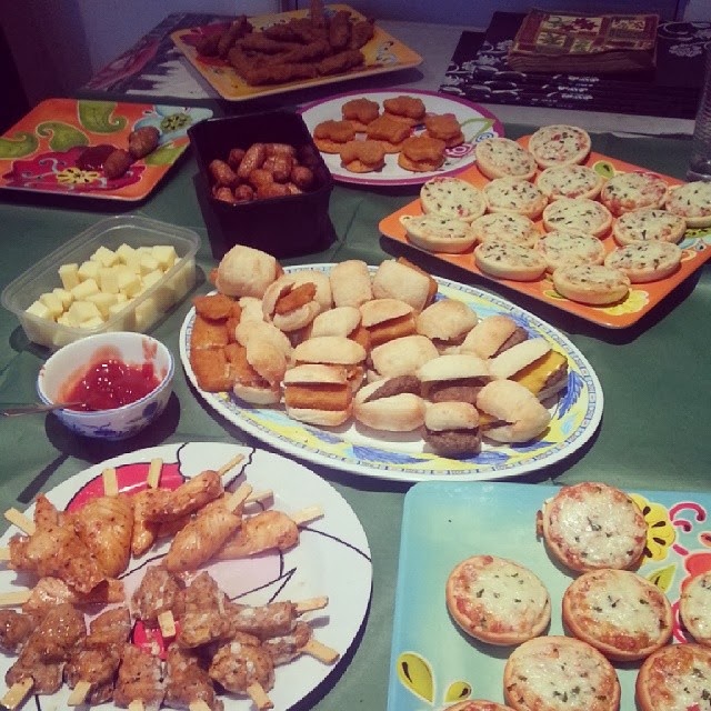 A table covered with various party foods, including mini pizzas, sliders, chicken skewers, cubes of cheese, breaded chicken goujons, sausage rolls, and bowls of dipping sauces, all arranged on colourful plates and trays.