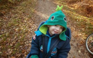 A young child wearing a dinosaur-themed green hood and blue jacket sits in a carrier on a forest path covered in fallen leaves. The child smiles at the camera. Part of a bicycle wheel and a dog walking ahead are visible in the background.