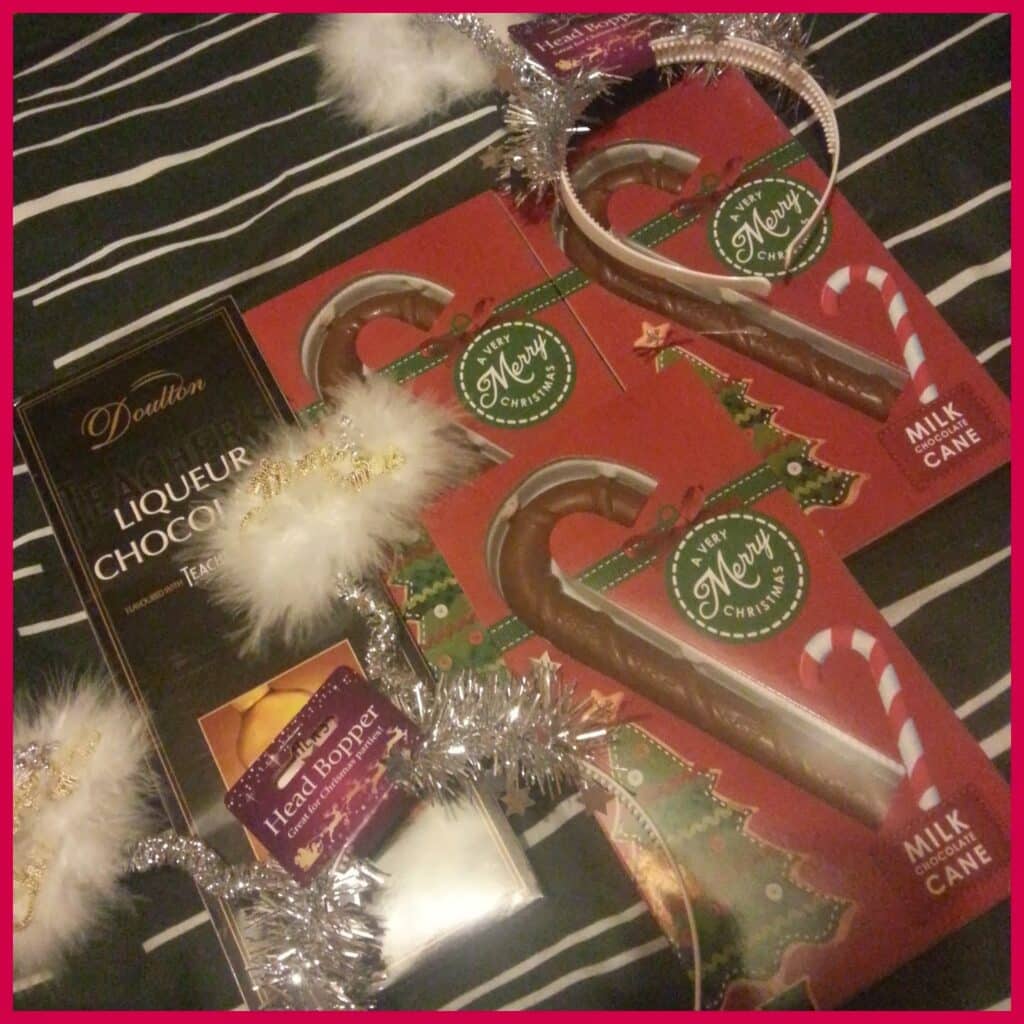 A photo shows two boxes of Merry Christmas milk chocolate candy canes, a Doalton liqueur chocolate bar, and two headbands with fluffy white halos and silver tinsel, all placed on a black and white striped fabric background.