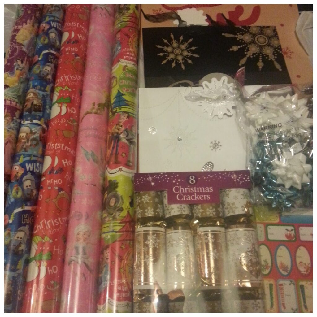 Several colourful rolls of Christmas wrapping paper are laid out beside white cards with silver snowflake designs, a black card with gold snowflakes, silver and gold Christmas crackers, gift bows, ribbon, and sheets of Christmas gift tags.