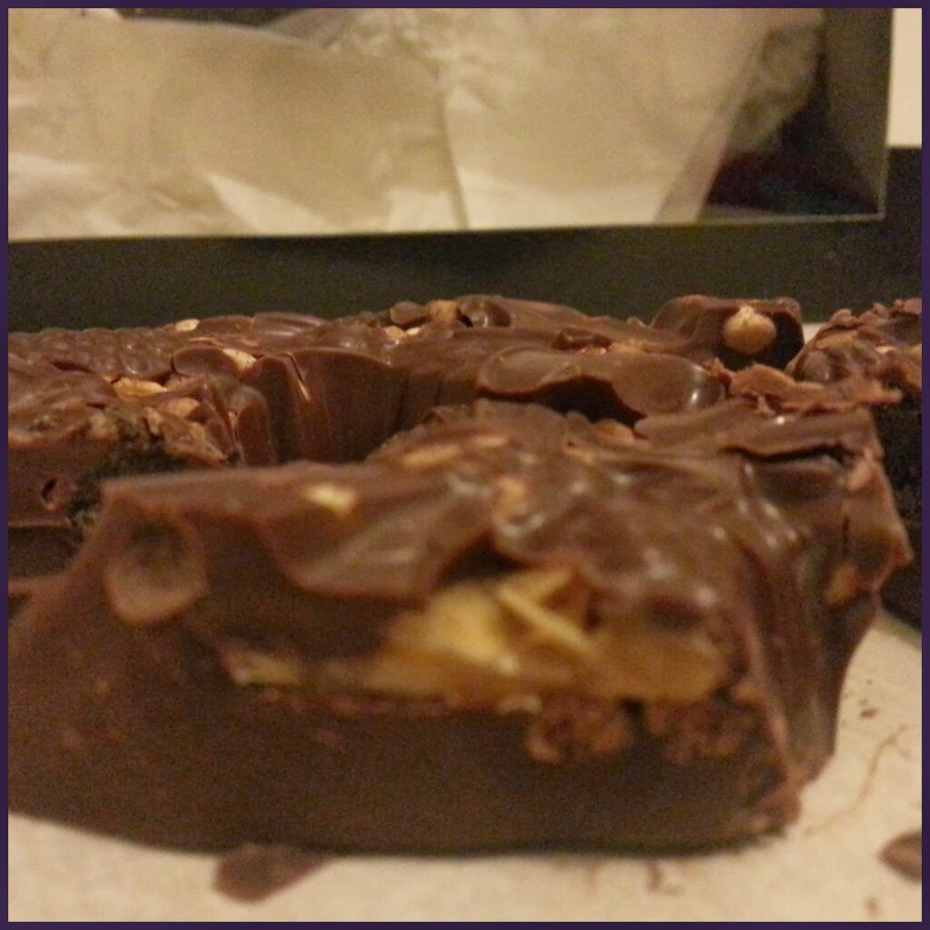 Close-up of a thick slice of chocolate with visible nuts embedded inside. The chocolate is glossy and chunky, with a textured surface. The background is blurry, showing a black box and white paper, suggesting the chocolate may be part of a gift or parcel.