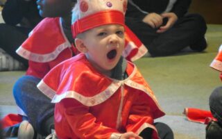 A young child in a red and white costume with a crown sits cross-legged on the floor, yawning widely. Other children in similar costumes are seated nearby, all indoors. The child’s facial expression suggests tiredness or boredom.