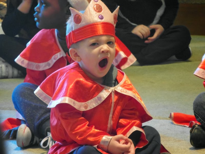 A young child in a red and white costume with a crown sits cross-legged on the floor, yawning widely. Other children in similar costumes are seated nearby, all indoors. The child’s facial expression suggests tiredness or boredom.