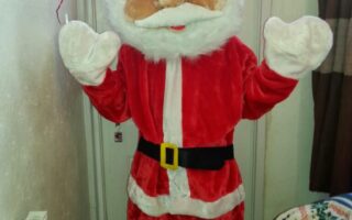 A person is wearing a plush Father Christmas costume with a large, round, cartoon-like head, white beard, red suit, black belt, and gloves. They stand indoors by a door, with one hand raised, next to a radiator and festive decorations.