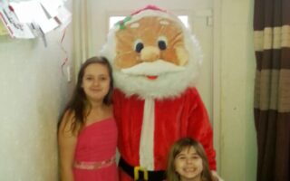 Three children pose indoors with a person in a large Father Christmas costume. Two girls wear pink and purple dresses, and a younger boy kneels in front wearing a grey shirt. They all smile, standing near a window with cards hanging on the wall.