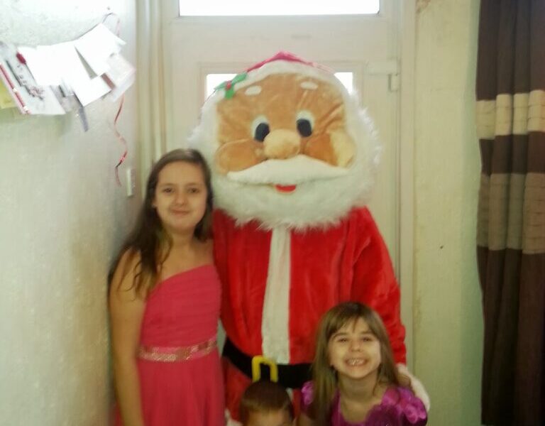 Three children pose indoors with a person in a large Father Christmas costume. Two girls wear pink and purple dresses, and a younger boy kneels in front wearing a grey shirt. They all smile, standing near a window with cards hanging on the wall.