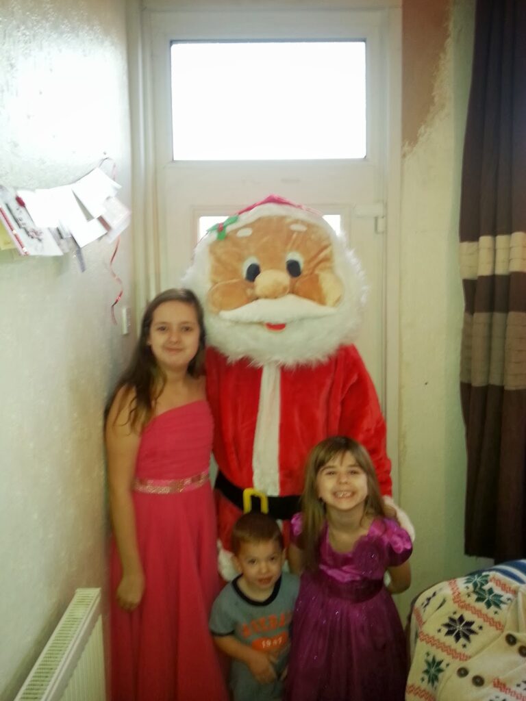 Three children stand indoors, smiling in front of a person dressed in a large Father Christmas costume. Two girls wear pink dresses, and a younger boy wears a grey T-shirt. They are near a white door, with cards hanging on the wall and brown curtains to the right.