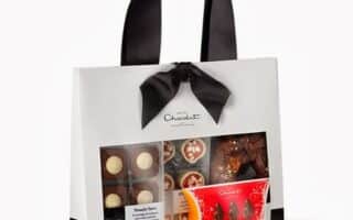 A gift bag with a black ribbon handle and bow, featuring a clear window displaying assorted chocolates in different shapes and colours. The bag is white and black with the “Hotel Chocolat” logo, and the chocolates are neatly arranged in trays and a red box.