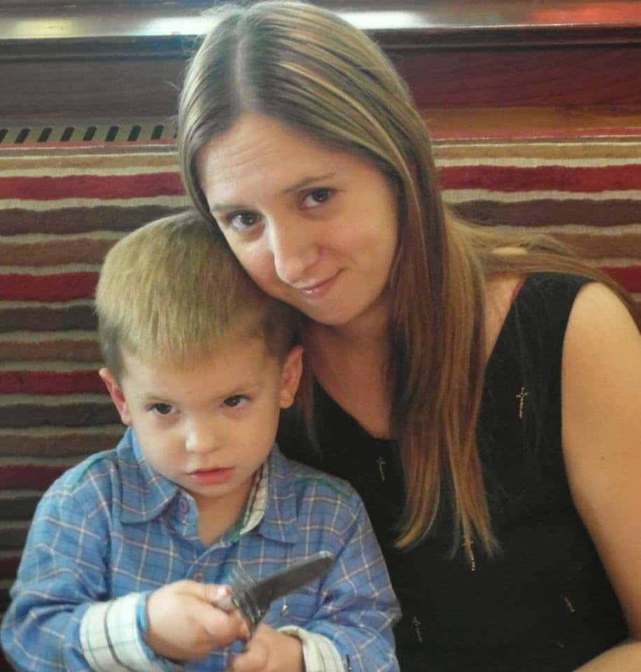 A woman with long brown hair in a black top sits closely beside a young boy in a blue checked shirt. The boy is holding a knife and looking ahead with a serious expression. They are seated on a striped upholstered bench.