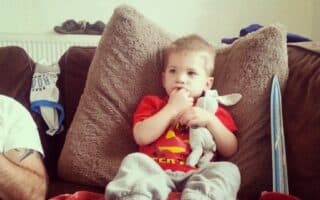 A young boy in a red shirt and grey tracksuit bottoms sits on a sofa against large brown cushions, holding a white soft toy and sucking his thumb. A blue toy sword, red cap, and another person’s arm are visible nearby.