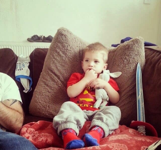 A young boy in a red shirt and grey tracksuit bottoms sits on a sofa against large brown cushions, holding a white soft toy and sucking his thumb. A blue toy sword, red cap, and another person’s arm are visible nearby.