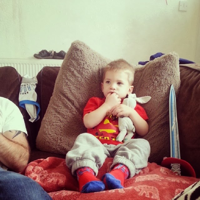 A young boy in a red shirt and grey tracksuit bottoms sits on a sofa against large brown cushions, holding a white soft toy and sucking his thumb. A blue toy sword, red cap, and another person’s arm are visible nearby.