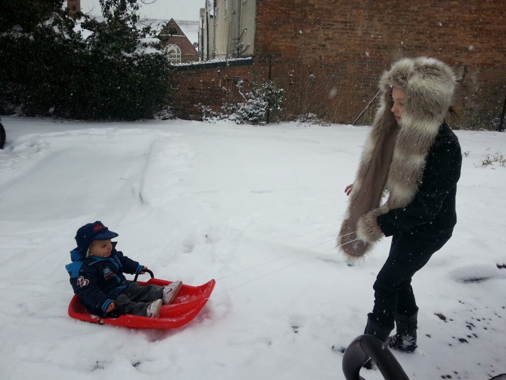A child in a black coat with a furry hood pulls a red sledge with a small child bundled in winter gear sitting on it. They are outside in a snow-covered garden with buildings and a brick wall visible in the background. Snow is falling gently around them.