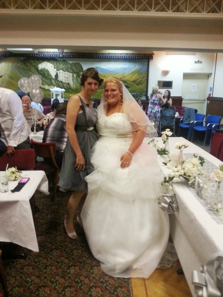 Two women pose together at a wedding reception. One wears a white wedding dress and veil, the other a grey dress. They are smiling in a decorated hall with white flowers, balloons, and seated guests. A mural is visible in the background.