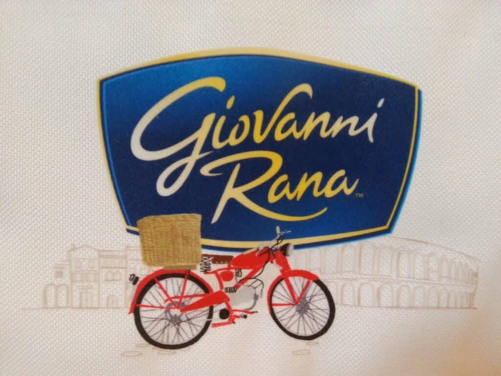 A blue and yellow Giovanni Rana logo appears above a red bicycle with a tan basket. The bicycle is illustrated on a white background with light line drawings of Italian architecture, including the Colosseum, in the background.