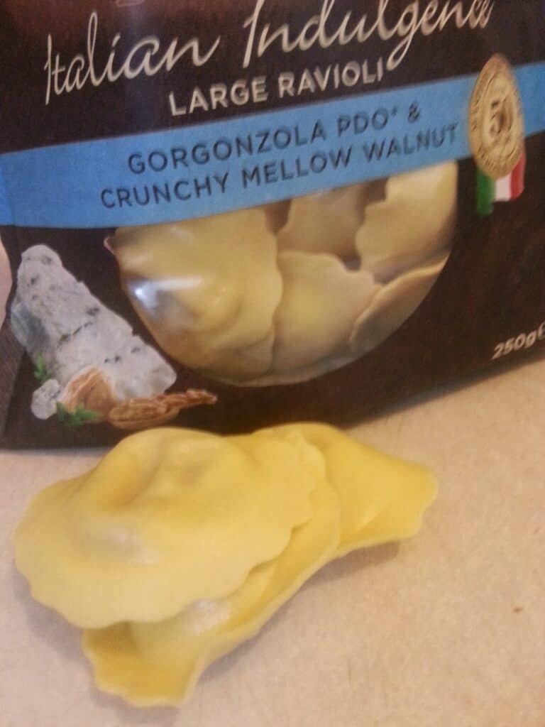 A package labelled "Italian Indulgence Large Ravioli—Gorgonzola PDO & Crunchy Mellow Walnut" stands upright, showing large, uncooked yellow ravioli inside. Three pieces of ravioli are placed outside the package on a beige surface.