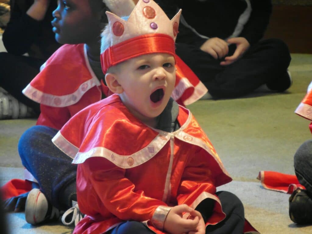 A young child wearing a red costume with a crown sits cross-legged on the floor, yawning. Other children in similar costumes sit nearby. The scene suggests a group activity or performance, and the child in focus appears tired or bored.