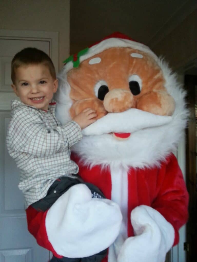 A smiling boy in a checked shirt is being held by a person dressed in a large, plush Father Christmas costume. The child touches Father Christmas’s fluffy white beard and looks at the camera, while the Father Christmas costume features a red suit and a big, friendly face.