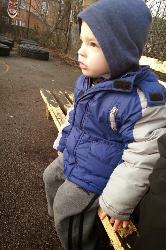 A young child in a blue and grey puffer jacket and grey tracksuit bottoms sits on a worn yellow wooden bench outdoors. He wears a blue hood and looks ahead seriously. Several stacked tyres and a fenced area are visible in the background.