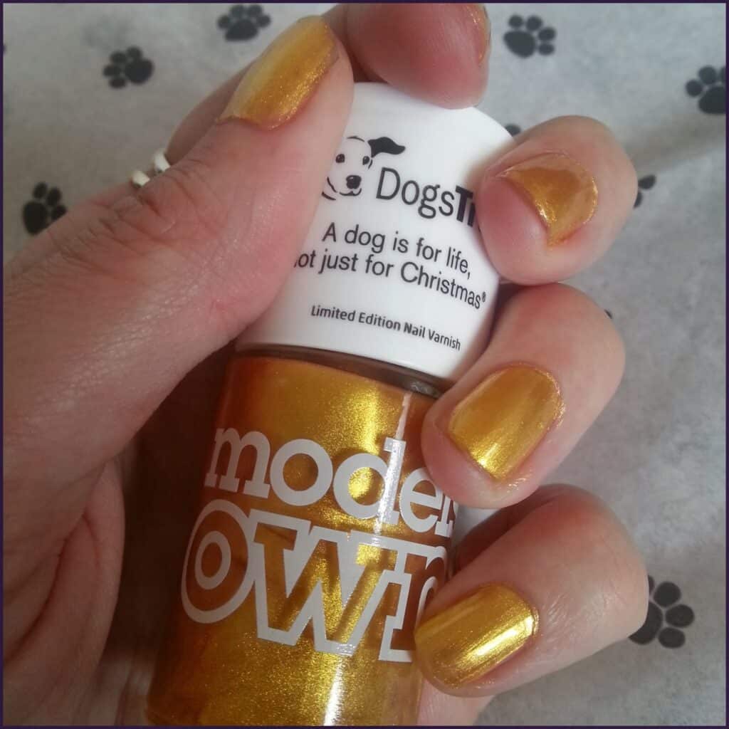 A hand with short nails painted metallic gold holds a matching gold nail varnish bottle labelled "models OWN." The bottle is topped with a white cap with black text and a dog logo. The background is light grey with faint black paw print patterns.