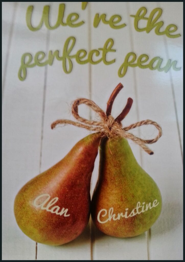 Two pears sit side by side on a wooden surface, tied together at the stems with string. Each pear has a name written on it: "Alan" and "Christine." Above them, green text reads, "We're the perfect pair.