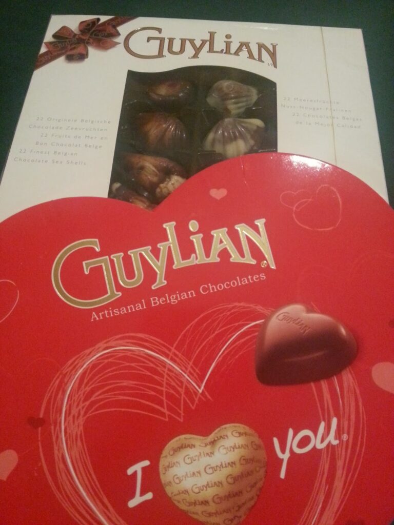 Two boxes of Guylian Belgian chocolates. The top box is rectangular with a window showing seashell-shaped chocolates. The bottom box is heart-shaped, red, with gold letters reading "Guylian" and “I love you” alongside a gold and red heart.