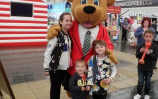 A person in a large bear costume wearing a red suit and tie poses with three smiling children. They stand on a paved area near a striped stall, with shops and people in the background. The scene appears to be at a lively indoor or covered event.