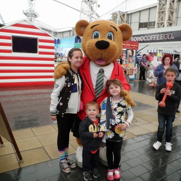 A person in a large bear costume wearing a red suit and tie poses with three smiling children. They stand on a paved area near a striped stall, with shops and people in the background. The scene appears to be at a lively indoor or covered event.