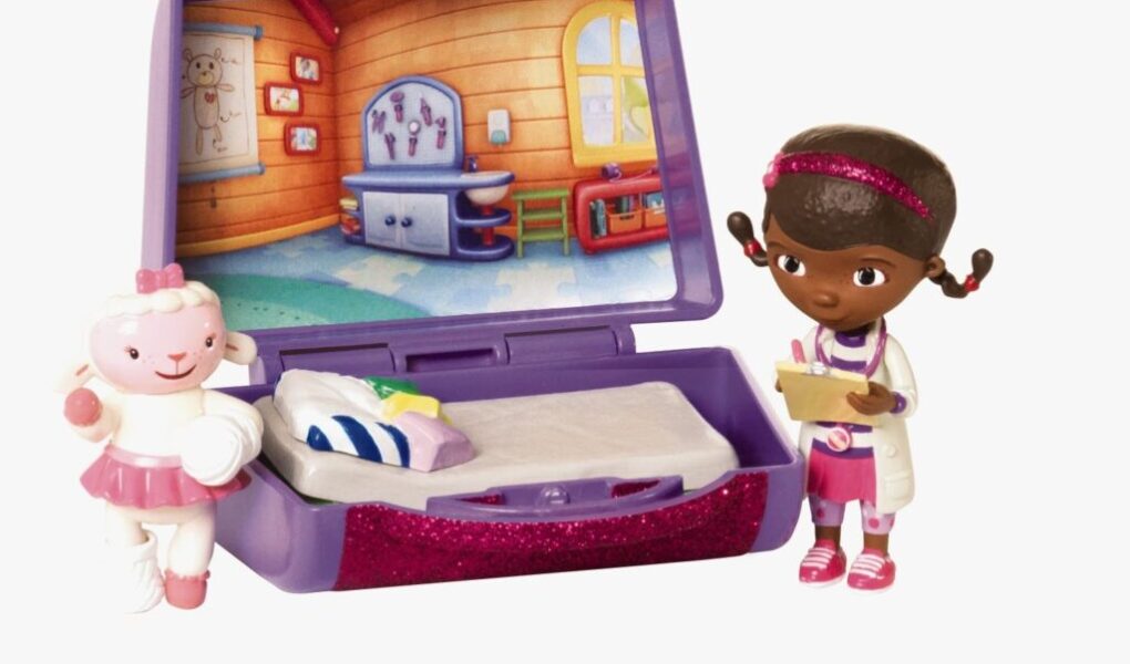 A toy doctor playset featuring a purple medical kit with a bed and a colourful clinic backdrop. A small figure of a girl in a doctor's coat holds a clipboard. Next to her is a sheep character wearing a pink tutu and bow, standing by the open kit.