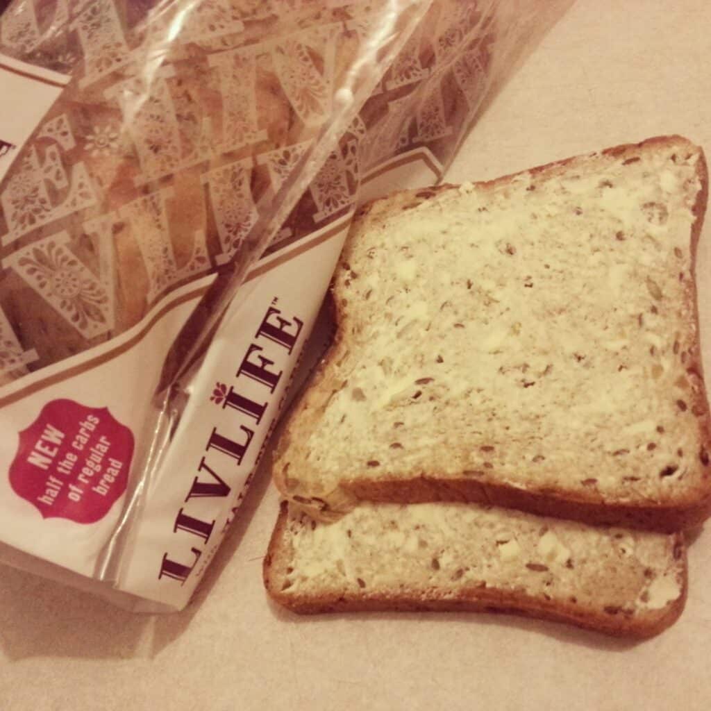 Two slices of multigrain bread, each spread with butter, rest on a light worktop. Next to them is their partially opened bread bag labelled “LIVLIFE.” The bread has visible seeds and grains; the packaging promotes it as a healthier bread choice.