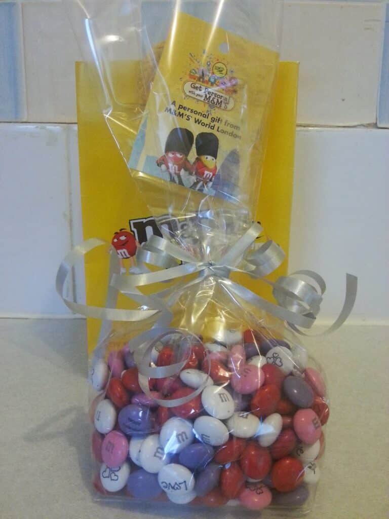A clear gift bag filled with red, pink, white, and purple M&M's, tied with silver ribbon, stands in front of a yellow M&M's branded bag. A gift card with cartoon M&M's characters is attached to the ribbon. The background is a tiled wall.