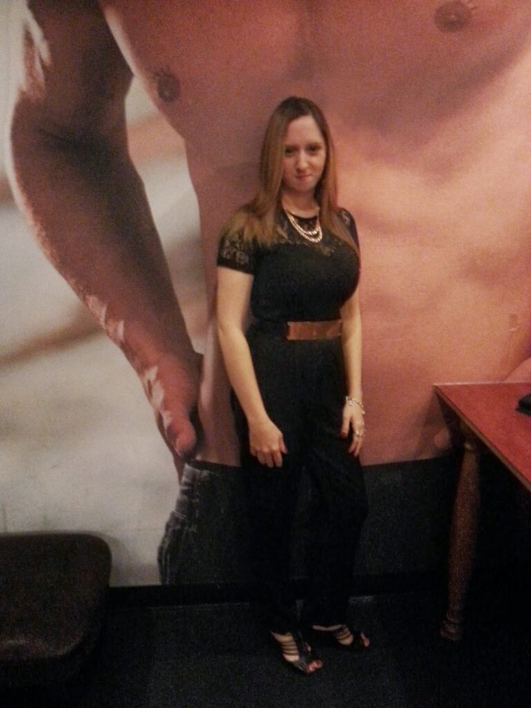 A woman with straight, light brown hair stands indoors against a wall featuring a large, shirtless male torso mural. She wears a black jumpsuit, brown belt, layered necklaces, and sandals, with her left hand resting on a wooden table.