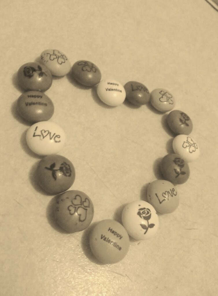 A heart shape made from sugar-coated chocolates lies on a light surface. Each sweet is decorated with words like “Love,” “Happy Valentine,” or simple drawings of roses and hearts. The image is in sepia tone, giving it a warm, nostalgic feel.