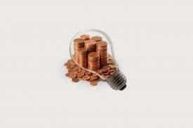 A lightbulb outline filled with stacked and scattered copper coins, symbolising the concept of saving money or financial ideas related to energy or innovation, set against a plain, light background.