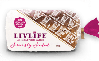 A loaf of LivLife Seriously Seeded bread is wrapped in white packaging with purple and pink text. The label highlights "half the carbs of regular bread," and the weight is 500g. A pink sticker seal is on the right side.