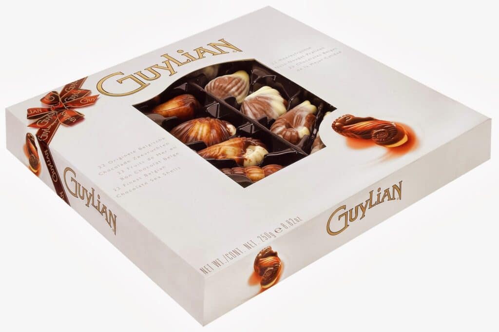 A box of Guylian Belgian chocolates with a clear window displays assorted seashell-shaped chocolates in marbled brown and white. The cream-coloured box features the Guylian logo in gold and images of the chocolates on the front and sides.