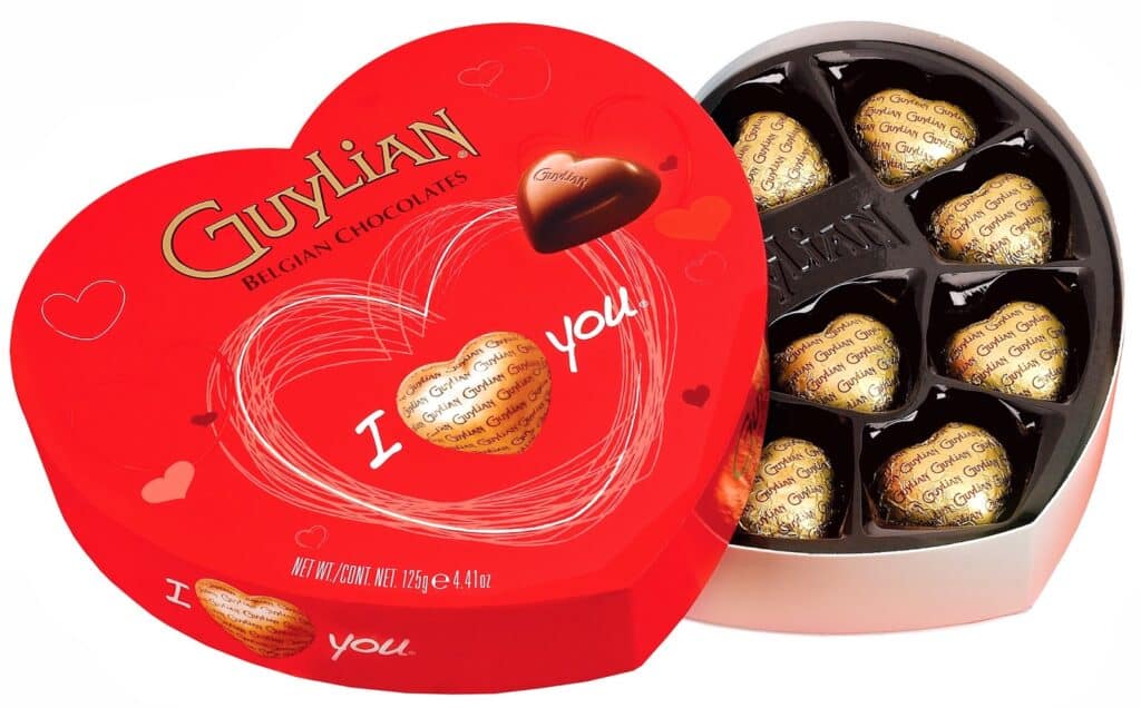 A red, heart-shaped box of Guylian Belgian chocolates with "I love you" and swirling heart designs on the lid. The box is open, revealing gold-foil-wrapped heart-shaped chocolates arranged neatly inside.