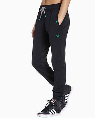 A person wears black Adidas jogger tracksuit bottoms with a green logo on the left pocket, green button details, and a grey drawstring. They pair the trousers with black Adidas trainers featuring white stripes and red accents. Their hands are in their pockets.