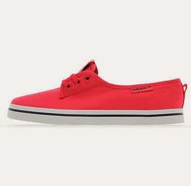 A bright red canvas trainer with a white rubber sole and thin black stripe along the side. The shoe has red laces, a low-cut design, and a small brand tag near the ankle. The background is plain and light-coloured.