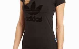 A woman with a plaited ponytail wears a fitted black Adidas t-shirt with a large black trefoil logo and “adidas” text on the front. She stands facing forwards, one arm relaxed at her side. Only her upper body is visible.
