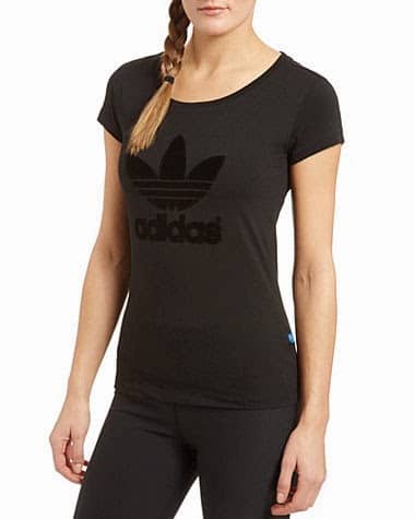 A woman with a plaited ponytail wears a fitted black Adidas t-shirt with a large black trefoil logo and “adidas” text on the front. She stands facing forwards, one arm relaxed at her side. Only her upper body is visible.